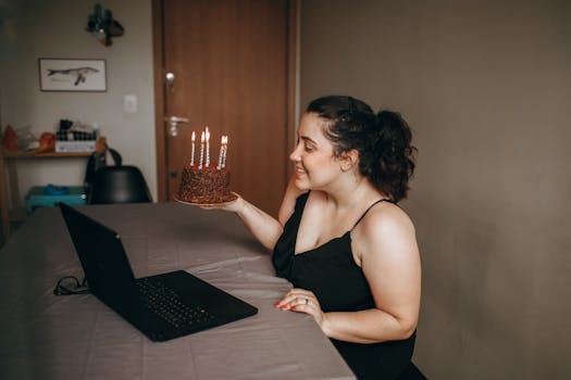 A woman enjoys a birthday celebration via video call with a chocolate cake and candles indoors.