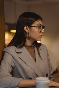 Side profile of a businesswoman wearing glasses and a blazer, sitting indoors in a cafe.